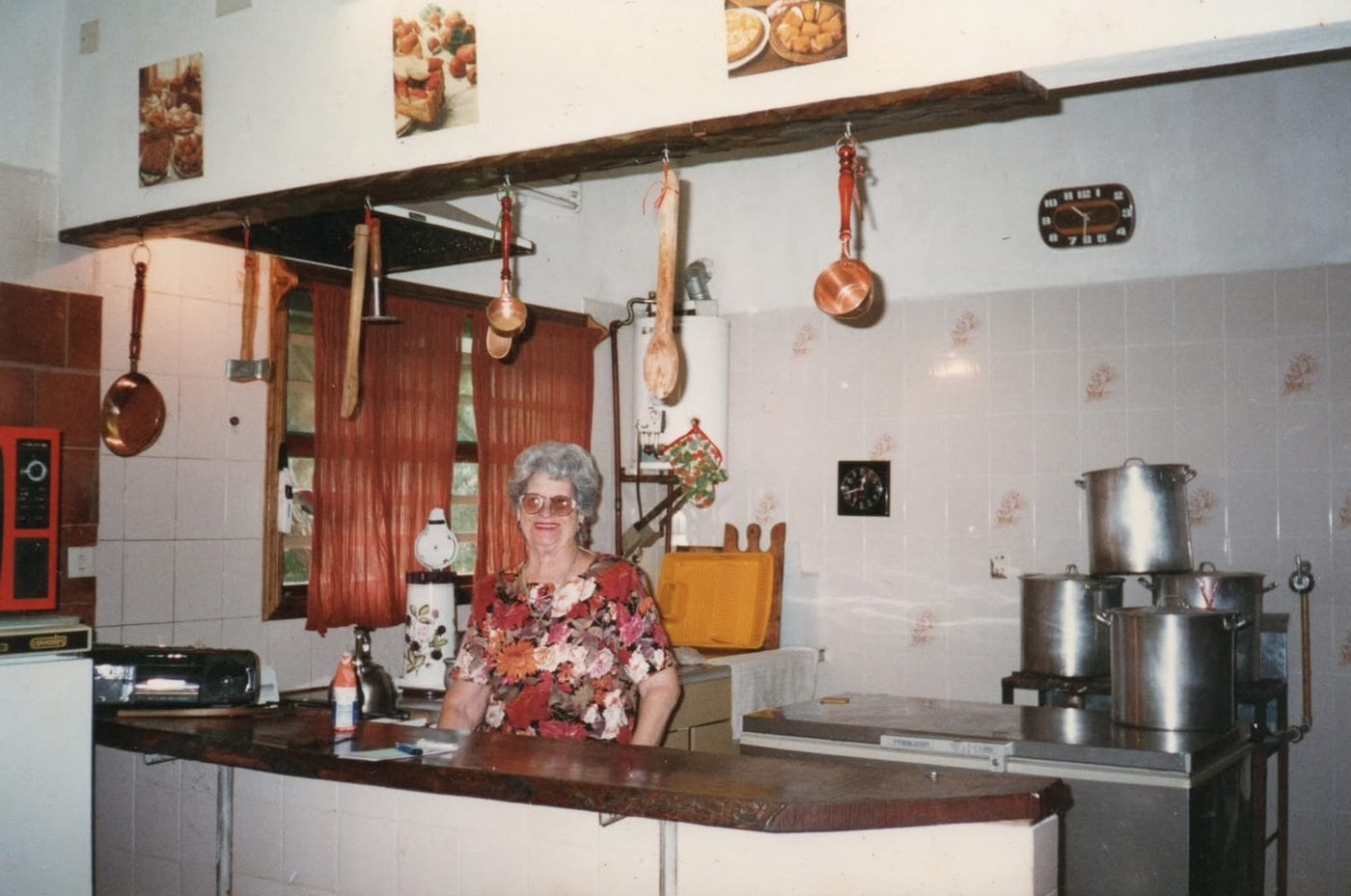 Foto antigua: una señora sonríe detrás de la mesada de una cocina con ollas grandes y utensilios de cobre colgados